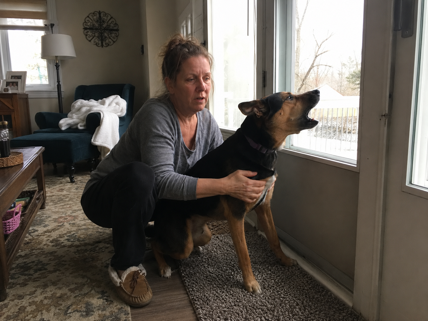 Owner crouched beside her dog at a window, mid-bark, looking overwhelmed — natural home setting
